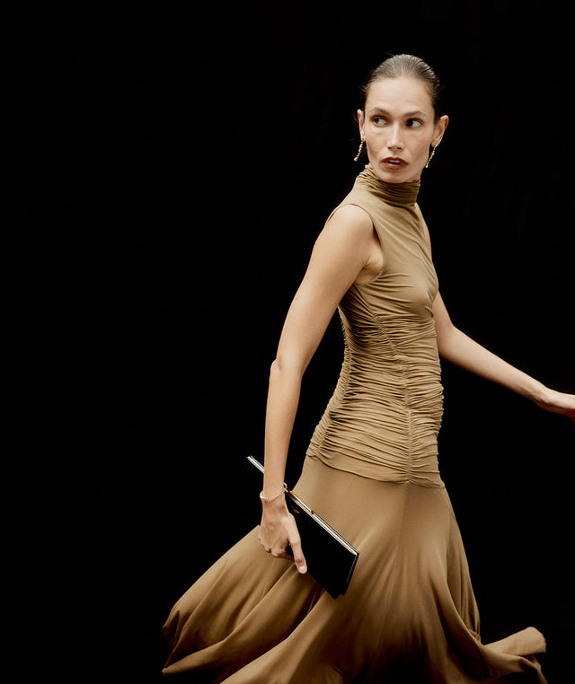 Woman in a brown Magda Butrym dress holding a Saint Laurent clutch against a black background