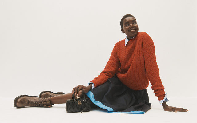 Woman wearing Miu Miu red sweater, navy pleated skirt, brown combat boos holding mini handbag