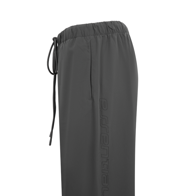 Image 3 of 3 - GREY - FEAR OF GOD ESSENTIALS Relaxed Track Pants featuring a drawstring waistband with long drawstrings, side hand pockets, wide leg openings with ankle zipper closures, seam details down the front legs, a silicone rubberized brand label on the front, a soft-touch Fear of God ESSENTIALS logo graphic down the leg and a baggy relaxed fit. 84% nylon, 16% spandex.