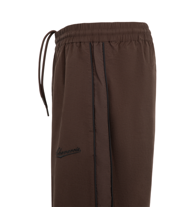 Image 3 of 3 - BROWN - Willy Chavarria Bad Boy Track Pant is crafted from lightweight nylon and features a relaxed athletic silhouette with an elasticated waistband and cuffs. Designed with a concealed drawstring waist, two seam pockets, embroidered logo detailing, and contrast piping along the outseams, the pant delivers the brand's bold sportswear-inspired aesthetic. Main Material: 100% Nylon.