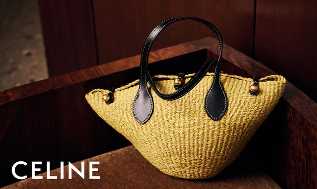 A Celine woven straw handbag in a warm yellow-gold tone with black leather handles and trim, resting on a dark wooden surface. The Celine brand name is displayed in white text in the bottom-left corner.