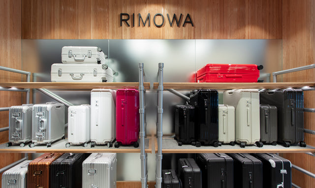 A retail display of RIMOWA hard-shell suitcases arranged on wooden shelves against a wood-paneled wall. Multiple colors are shown, including silver, white, red, black, beige, and brown.