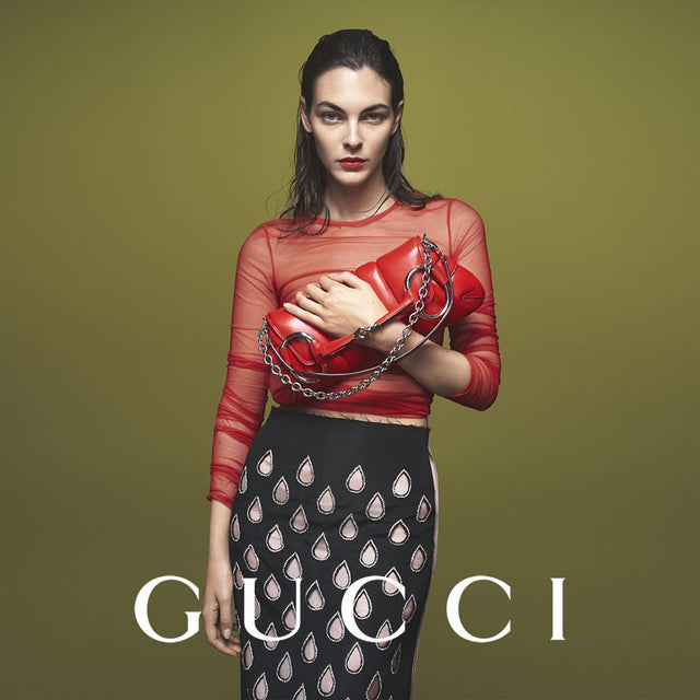 Gucci campaign- model wearing red long-sleeve top, black tear drop pattern skirt and holding red leather handbag with silver-tone hardware, Available in-store only at HIRSHLEIFERS.
