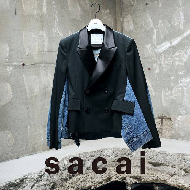 Black coat with denim sleeves on a hanger against a concrete wall, featuring the sacai brand. 