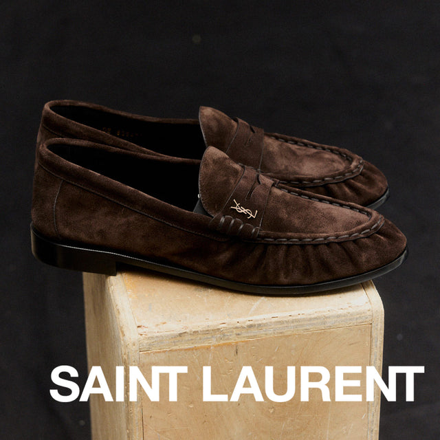 Brown suede loafers on a wooden block with 'Saint Laurent' branding against a dark background. 