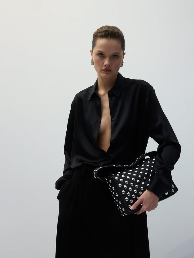 woman wearing black button up blouse and holding black leather handbags with silver studs, both by Khaite 
