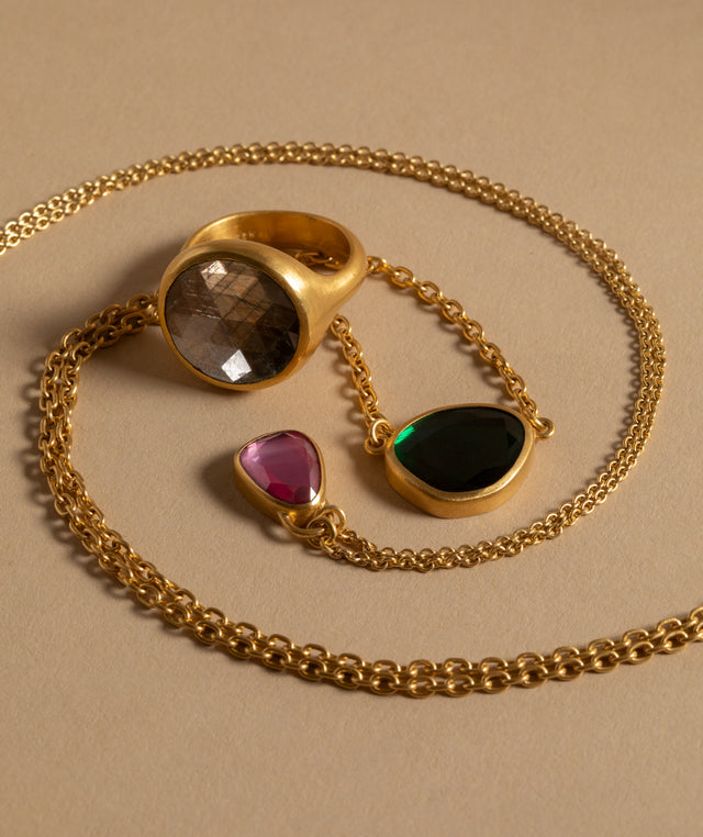 Eli Halili Gold chain jewelry - gold ring with a faceted smoky brown sapphire, a teardrop pendant with a dark green stone, and a small pendant with a pink ruby, arranged on a beige background. 