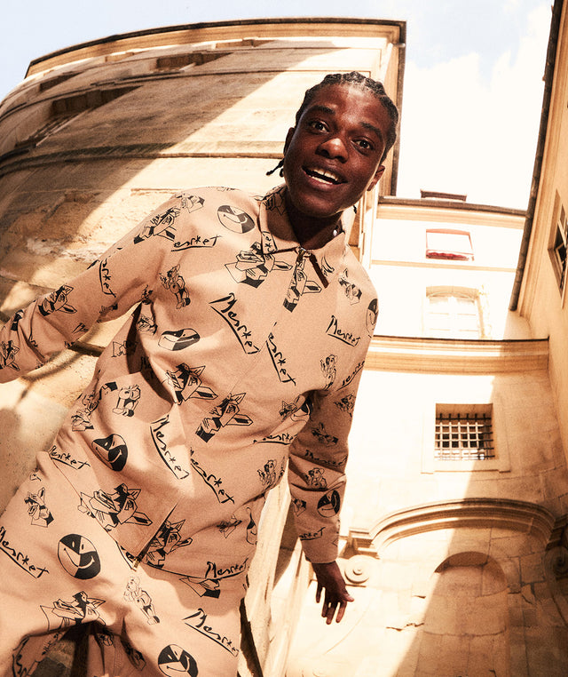 Person wearing a Market Studios smiley patterned shirt standing in front of a building with large windows. 