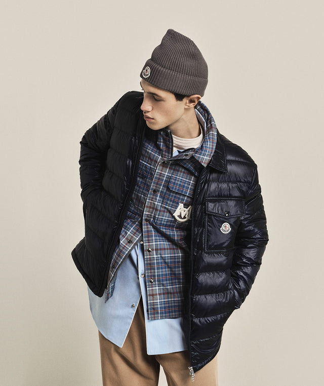 Person wearing a black Moncler jacket, plaid scarf, and beanie on a beige background 