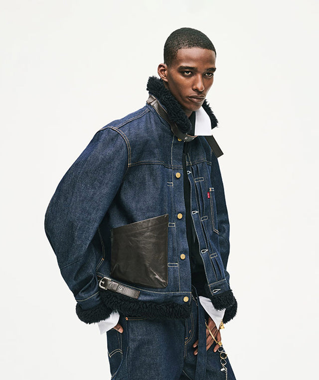 A male model wearing Lev's x sacai dark blue denim jacket with black shearling lining, a black leather chest patch pocket, and a matching denim jacket and jeans 