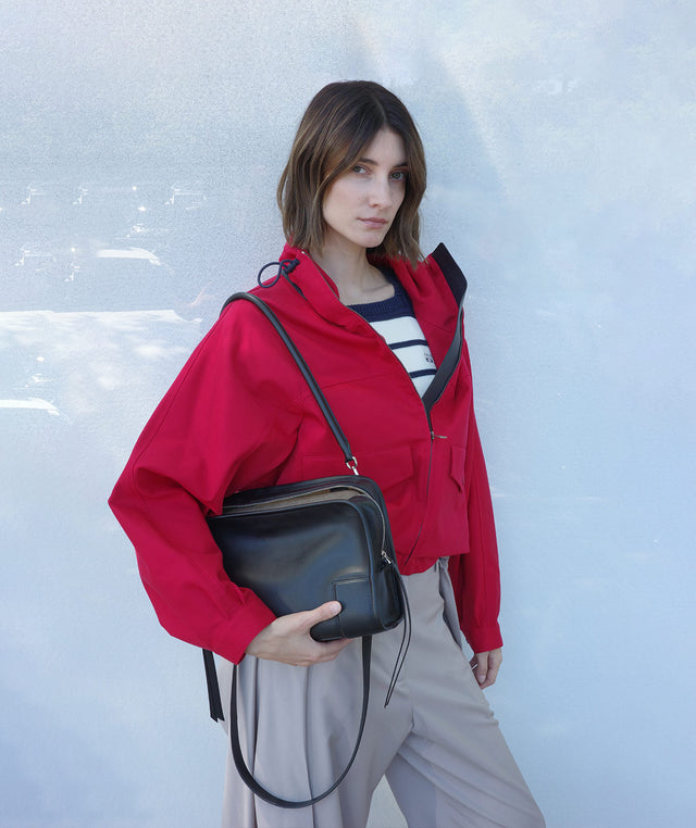Woman in a red jacket by Loewe holding a black Amazona 180 handbag against a light blue wall. 