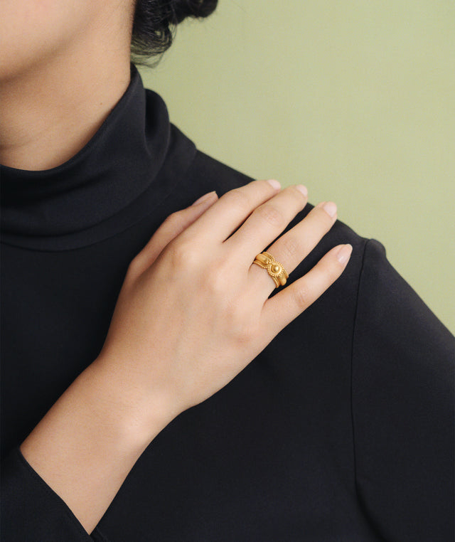 A close-up of a person wearing a textured gold Prounis ring on their hand, dressed in a black turtleneck against a soft light green background. 