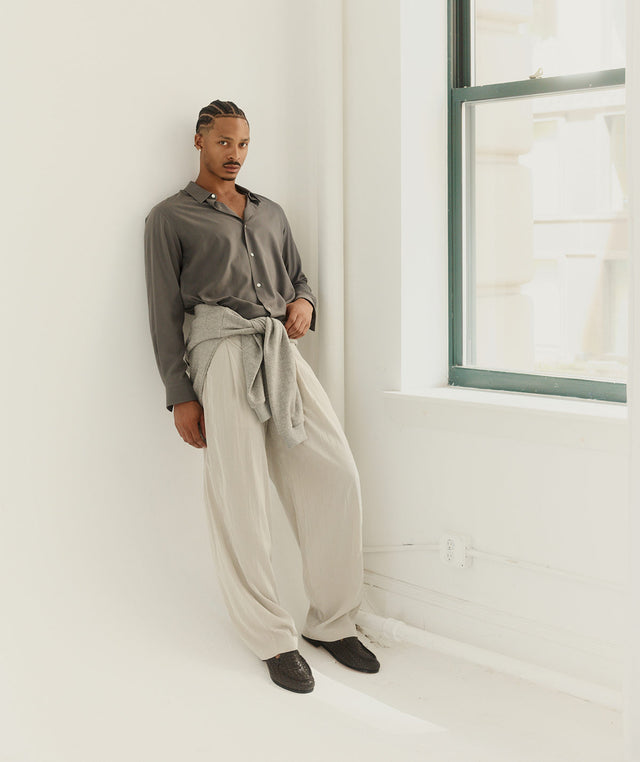 Person wearing a gray shirt and beige pants by The Row standing in a minimalistic room with a window. 