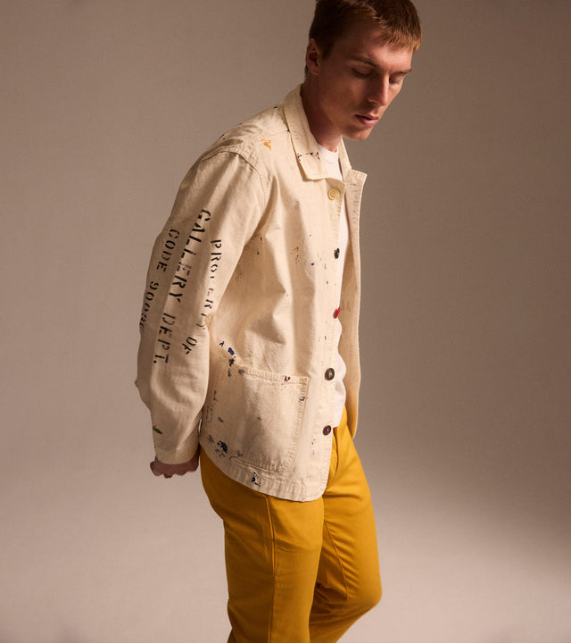 Man wearing white denim jacket splattered with paint and yellow chino pants, both by Gallery Dept. 