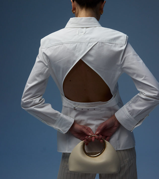 Woman wearing JACQUEMUS Open Back Shirt made of white cotton poplin and holding JACQUEMUS Le petit Calino Bag crafted from creme nappa lambskin with fixed metal carry handle