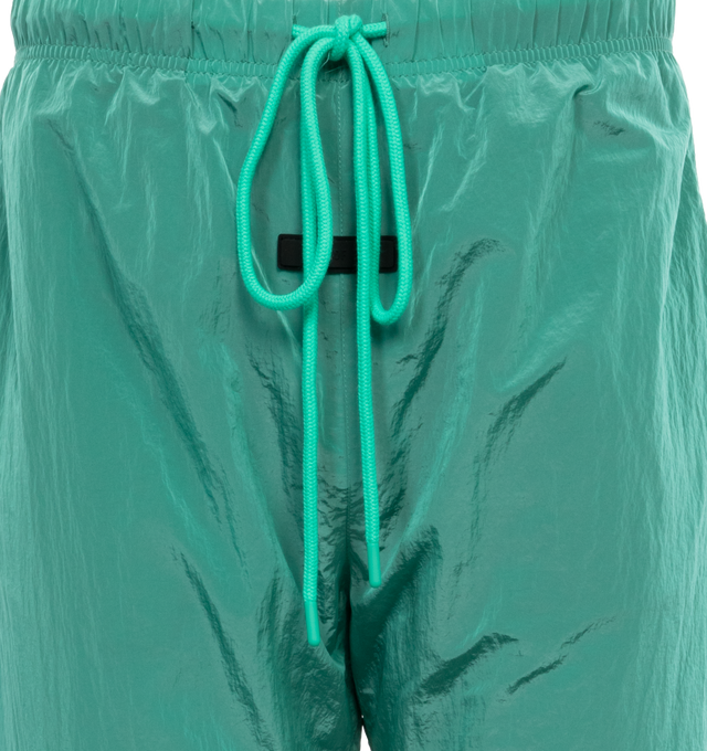 Image 4 of 4 - GREEN - FEAR OF GOD ESSENTIALS Crinkle Nylon Trackpants featuring an encased elastic waistband with elongated drawstrings, side seam pockets, an elastic hem with zipper adjustability at the ankle and a rubberized label at the center front. 100% nylon.  