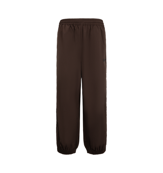 Image 1 of 3 - BROWN - Willy Chavarria Bad Boy Track Pant is crafted from lightweight nylon and features a relaxed athletic silhouette with an elasticated waistband and cuffs. Designed with a concealed drawstring waist, two seam pockets, embroidered logo detailing, and contrast piping along the outseams, the pant delivers the brand's bold sportswear-inspired aesthetic. Main Material: 100% Nylon.  