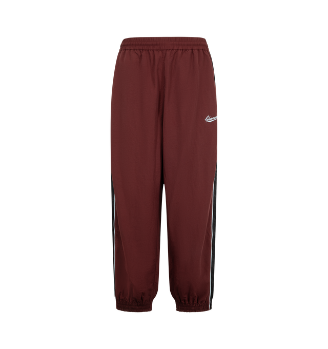 Image 1 of 3 - RED - Willy Chavarria Bad Boy Track Pant is crafted from lightweight nylon and features a relaxed athletic silhouette with an elasticated waistband and cuffs. Designed with a concealed drawstring waist, two seam pockets, embroidered logo detailing, and contrast piping along the outseams, the pant delivers the brand's bold sportswear-inspired aesthetic. Main Material: 100% Nylon.  