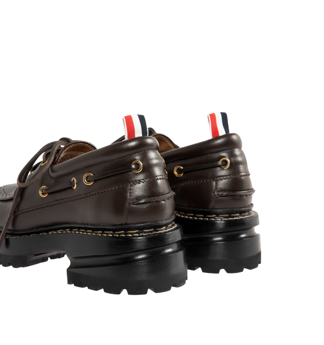 Image 3 of 4 - BROWN - THOM BROWNE Vitello Lug Boat Shoes featuring grained calfskin boat shoes, bumper hardware at toe with enameled stripes, perforated detailing at moc toe, lace-up closure, padded collar, serrated edge at sides and heel counter, tricolor grosgrain pull-loop at heel collar, buffed calfskin lining, stacked leather midsole and heel and treaded rubber sole. Heel: H2". Calfskin. Sole: rubber. Made in Italy. 