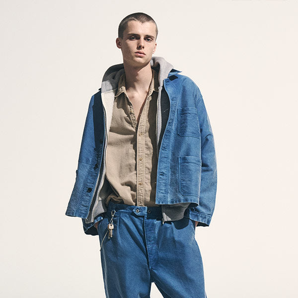 A male model wearing a blue denim jacket over a beige button-up shirt and blue jeans all in A.PRESSE.