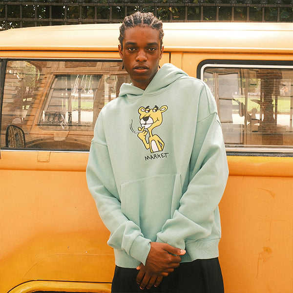 Person wearing a Market Studios light blue hoodie with a cartoon panther design, standing in front of a yellow vehicle.