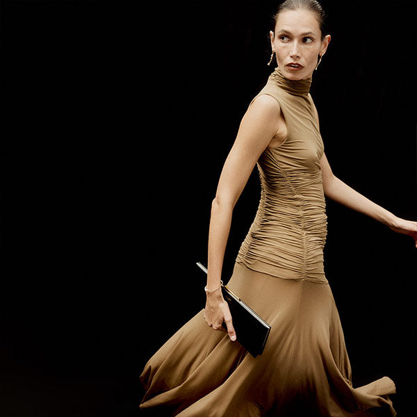 Woman in a brown Magda Butrym dress holding a Saint Laurent clutch against a black background