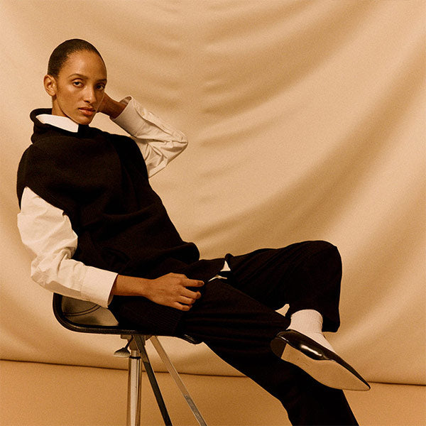 Person sitting on a chair wearing a black and white outfit by The Row against a beige background