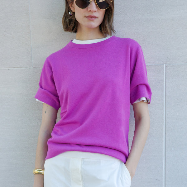Person wearing a bright pink sweater by Extreme Cashmere over a white shirt with white pants, standing against a light gray wall.