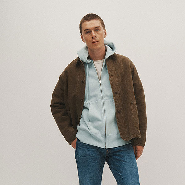 Man wearing Kaptain Sunshine brown workwear jacket, light blue hooded sweatshirt and denim jeans