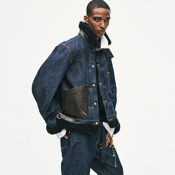 A male model wearing Lev's x sacai dark blue denim jacket with black shearling lining, a black leather chest patch pocket, and a matching denim jacket and jeans