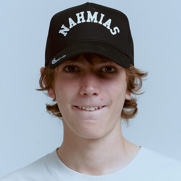 A young man smiling while wearing a black trucker-style cap with 'NAHMIAS' in white arched lettering on the front, set against a solid light blue background.
