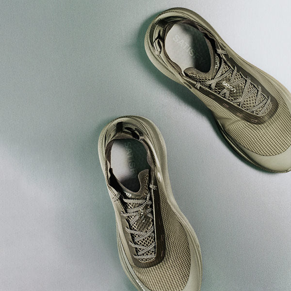A pair of Satisfy athletic sneakers in a green colorway, photographed from a top-down angle on a smooth, light gray surface.