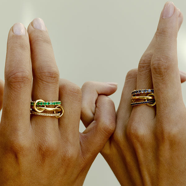 Two hands wearing Spinelli Kilcollin gold linked rings with emeralds and diamonds