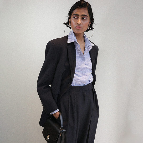 Women wearing black blazer, blue and white striped button-up shirt, black pleated dress pants and holding a black leather top-handle purse, all by The Row