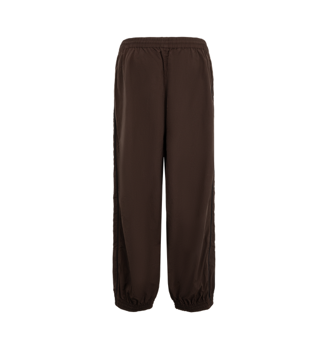 Image 2 of 3 - BROWN - Willy Chavarria Bad Boy Track Pant is crafted from lightweight nylon and features a relaxed athletic silhouette with an elasticated waistband and cuffs. Designed with a concealed drawstring waist, two seam pockets, embroidered logo detailing, and contrast piping along the outseams, the pant delivers the brand's bold sportswear-inspired aesthetic. Main Material: 100% Nylon.  