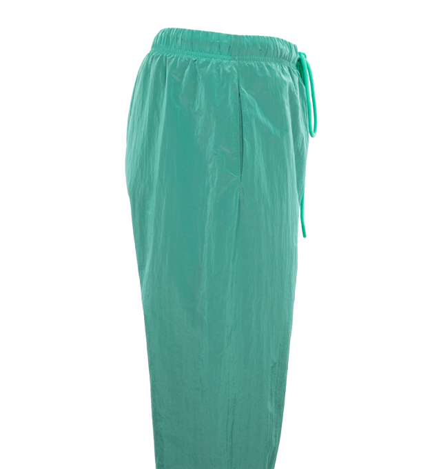 Image 3 of 4 - GREEN - FEAR OF GOD ESSENTIALS Crinkle Nylon Trackpants featuring an encased elastic waistband with elongated drawstrings, side seam pockets, an elastic hem with zipper adjustability at the ankle and a rubberized label at the center front. 100% nylon.  