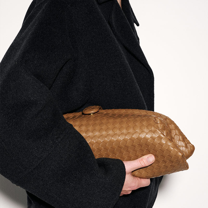 A person in a black coat holds a bottega veneta brown woven leather clutch bag with a gold-tone clasp against a plain light background.