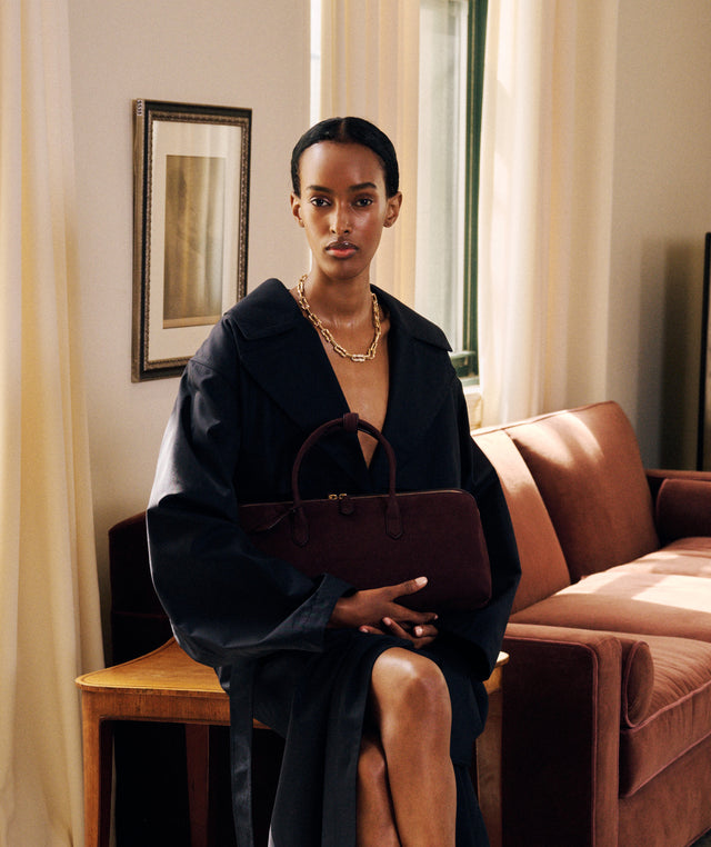 A model sits in a warm, sunlit living room holding a Alaia dark brown leather handbag and wearing black oversized trench coat by Alaia with a gold chain necklace. 