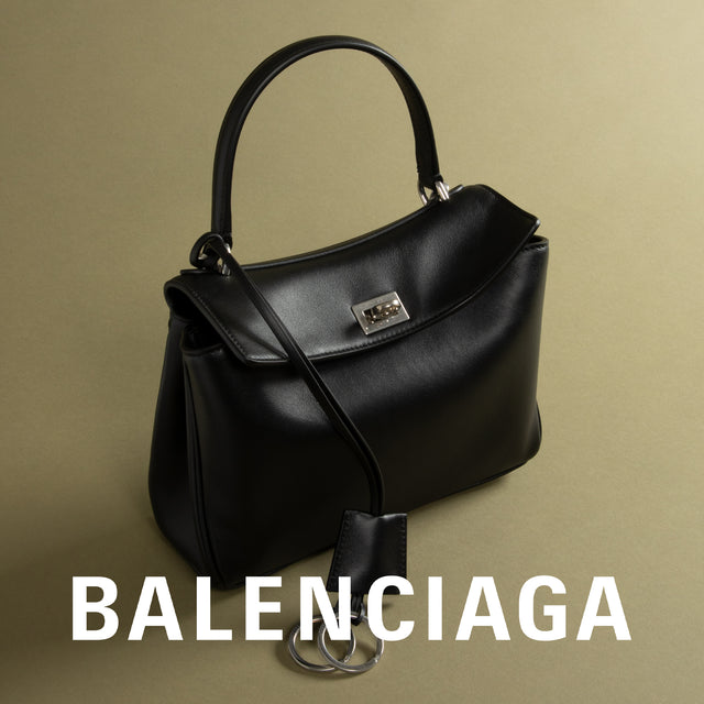 A black leather Balenciaga handbag with a top handle, a shoulder strap, and a silver-tone logo clasp, shown against a warm beige background with the Balenciaga brand name in white text at the bottom. 