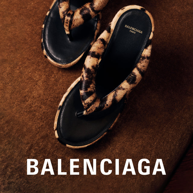 A pair of Balenciaga thong sandals with black leather soles and fuzzy leopard-print straps, displayed on a textured brown background with the Balenciaga brand name printed in white at the bottom. 