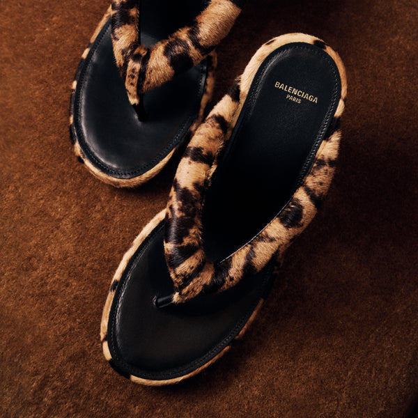 A pair of Balenciaga thong sandals with black leather straps and fuzzy leopard-print trim, photographed on a textured brown background.
