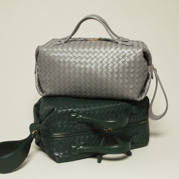 Two Bottega Veneta woven leather travel bags stacked on a light beige background: a silver top bag and a dark green bottom bag, both with top handles and shoulder straps.