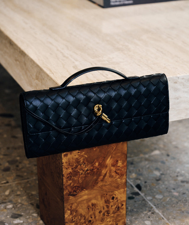 Bottega Veneta black woven leather clutch bag with a top handle and a gold key-shaped clasp, resting on a wooden block in front of a light wood table. 