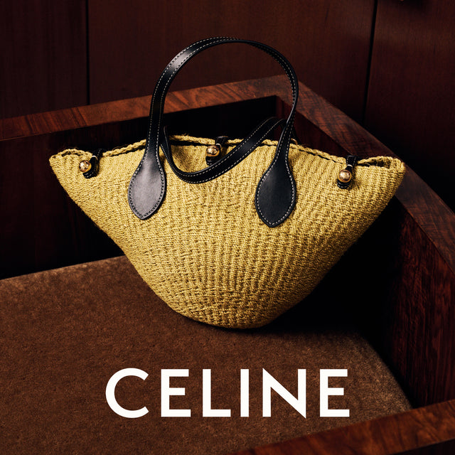 A woven straw handbag in a mustard yellow color with black leather handles and trim, displayed on a dark wooden bench. The Celine brand name is printed in white at the bottom of the image. 