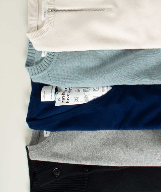Four folded knit crewneck sweaters stacked vertically, showing different colors: off-white, light blue, navy blue, and heather gray, with a black sweater partially visible at the bottom all in Extreme Cashmere brand. 
