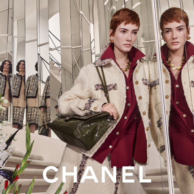 A model in front of mirrored walls wearing a Chanel outfit: cream bouclé jacket with ornate dark and metallic trim, a deep burgundy top, and a dark green crocodile-embossed handbag. 