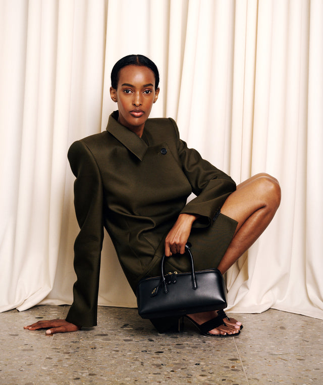 A model crouches in front of cream draped curtains, wearing a structured olive green suit with a high collar by Khaite and holding a black leather handbag. 