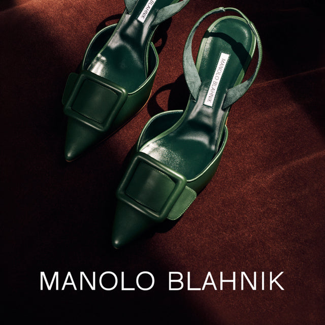 A pair of Manolo Blahnik pointed-toe slingback sandals in dark green leather with square buckle details, displayed on a dark brown textured surface with the brand name printed at the bottom. 