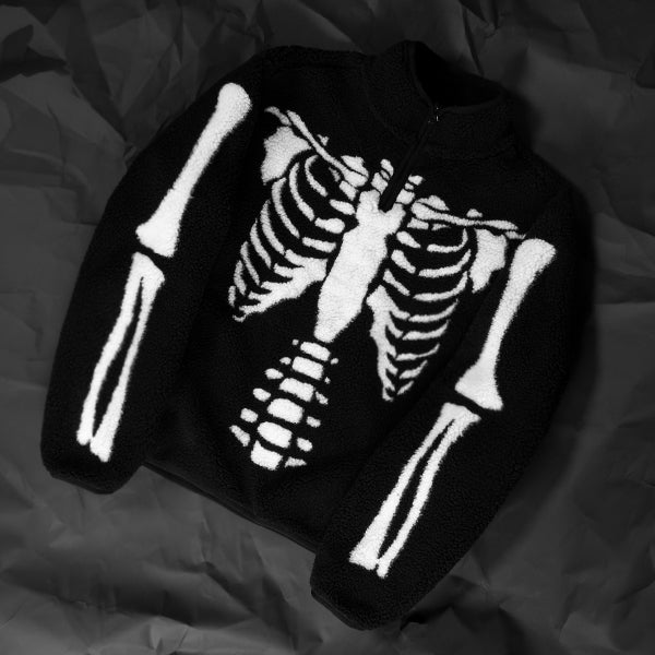 A black zip-up fleece sweater featuring a large white rib cage and arm bone graphic on the front, laid flat on a dark wrinkled fabric background.