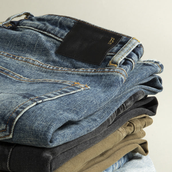 A close-up stack of folded denim jeans by R13 in different washes, with the top pair showing a black brand patch and gold lettering, photographed against a plain light background.